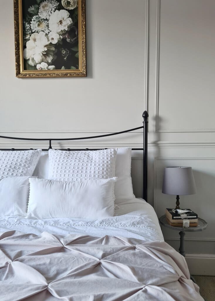 Image shows a bed with white bedding against a soft grey wall with wall panelling and framed oil painting. Side table with lamp and books. From a blog post about neutral paint colours by Life with Holly