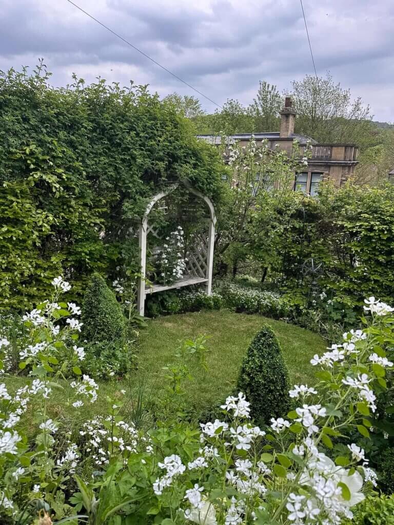 A lush green garden with white flowers and a white arbour partially hidden by a climbing rose